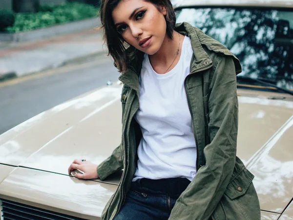 A woman in a white t-shirt and jacket sits on a car, smiling and enjoying the outdoors.