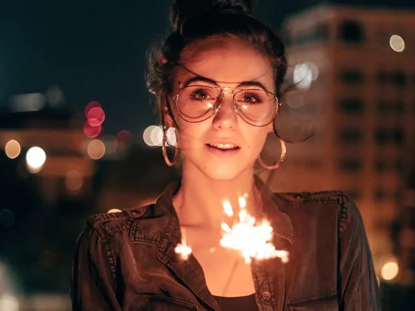 A woman with glasses joyfully holds sparklers, illuminating her face in the dark night sky.